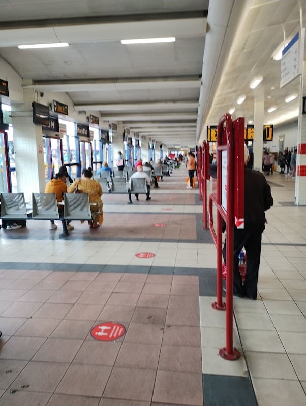 Covered bus station with seating areas, passengers waiting, and signage indicating bus routes in Huddersfield, Yorkshire.