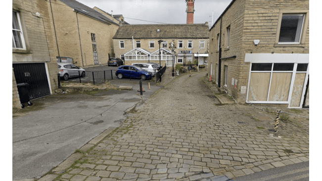 Cobblestone pathway leading to a parking area with cars, buildings, and a greenhouse in Huddersfield, Yorkshire.