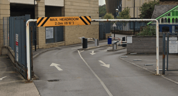 Pay & Display entrance with a height restriction sign, arrows directing traffic, and surrounding buildings in Huddersfield.