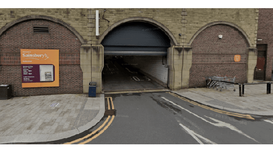 Entrance to a car park with a Sainsbury's sign, brick walls, and shopping trolleys nearby.