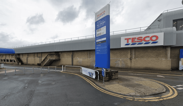 Tesco store exterior in Huddersfield with a large sign, parking area, and cloudy sky.