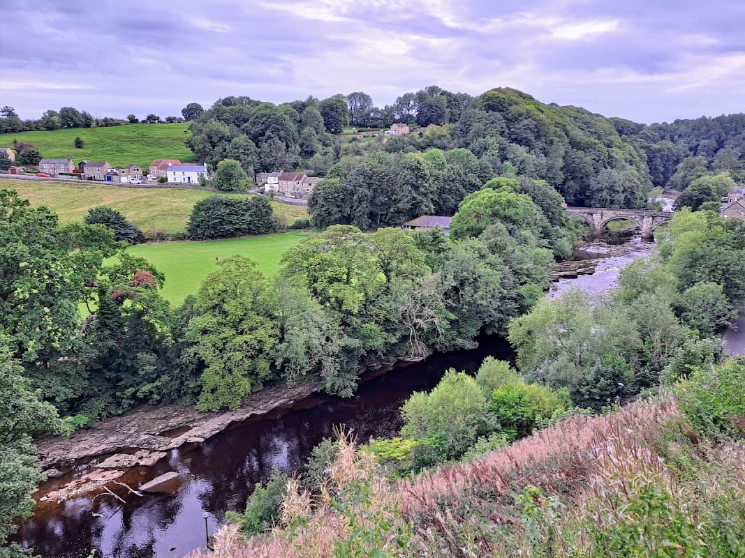 Lush green hills with trees, a river winding through, and quaint houses in Hudswell, North Yorkshire.