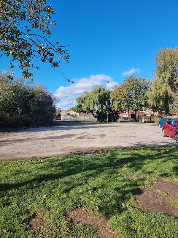 Alderman Kneeshaw Car Park with a clear blue sky, green grass, and trees surrounding a gravel parking area.