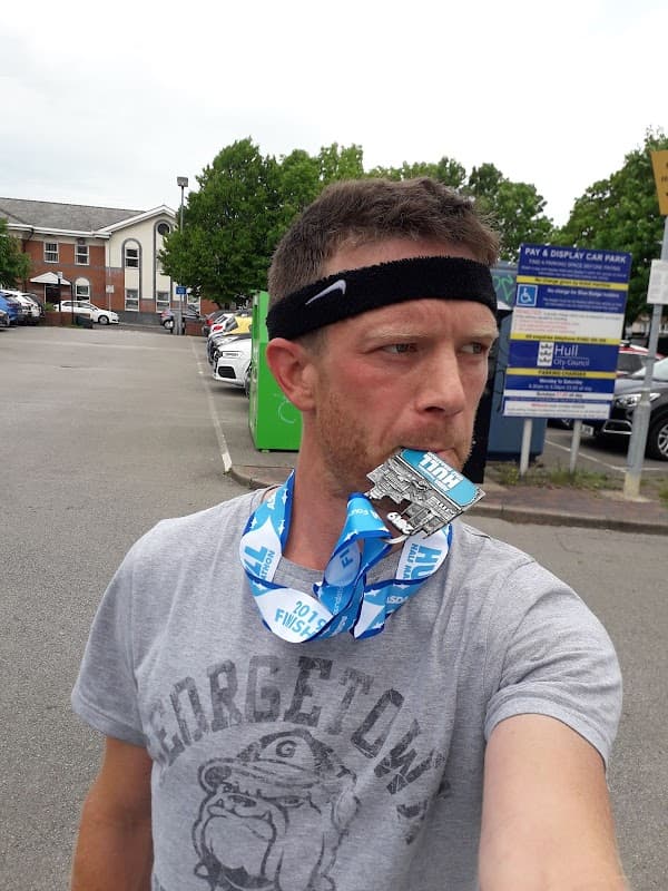 A man wearing a headband and a Georgetown t-shirt holds a medal in his mouth, standing in a car park.