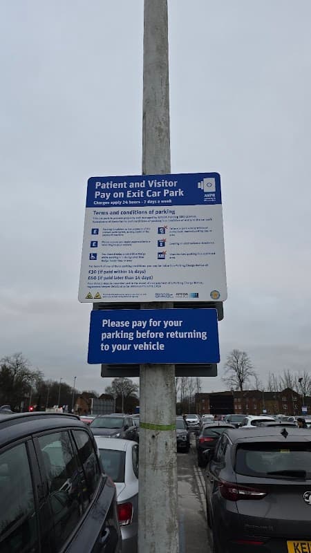 Signpost detailing parking terms and conditions in a car park filled with parked vehicles under a cloudy sky.