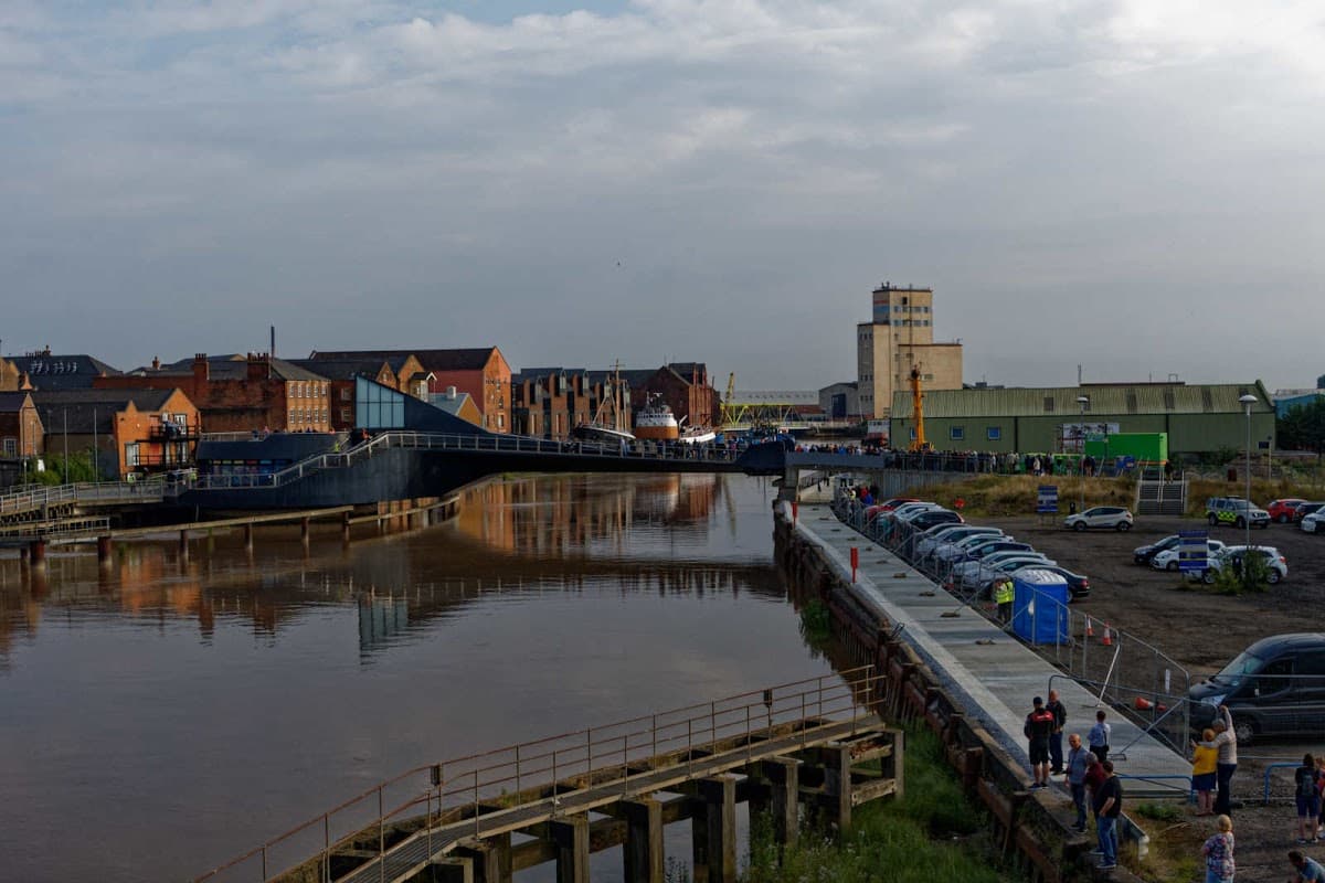 Pay & Display car park by the river, with parked cars, buildings, and a bridge in the background under a cloudy sky.