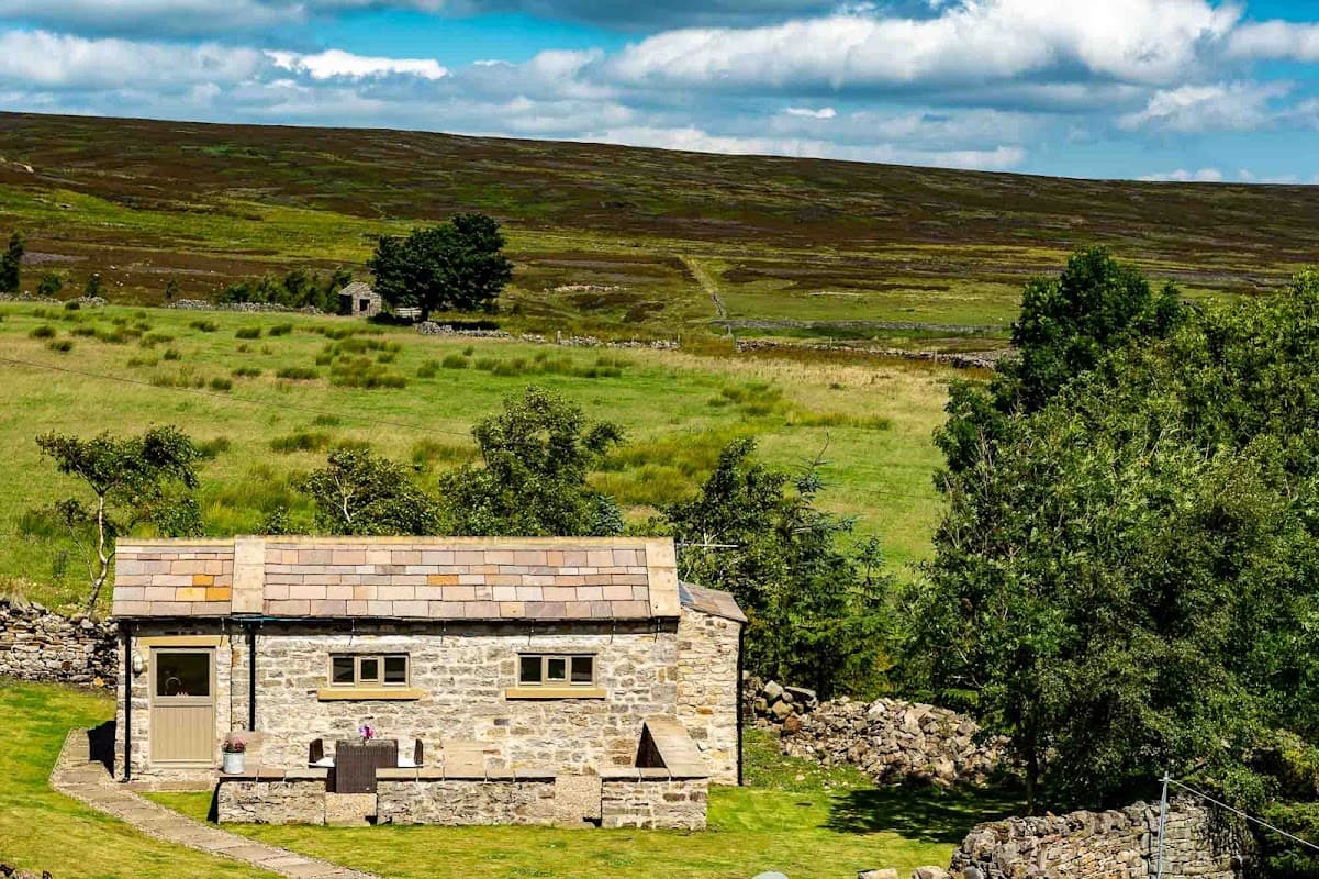 A stone cottage with a slate roof surrounded by green fields and hills under a blue sky with fluffy clouds.