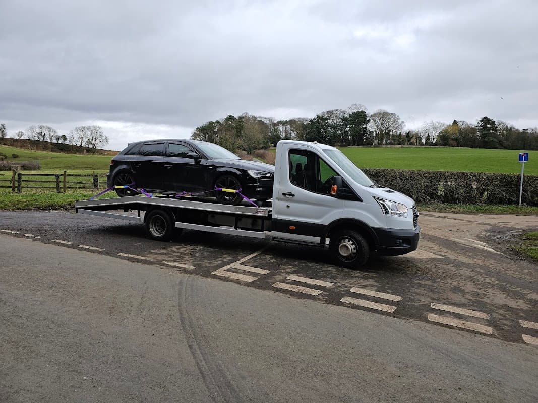 A flatbed truck with a black car secured on it, parked near a grassy field in Husthwaite, Yorkshire.