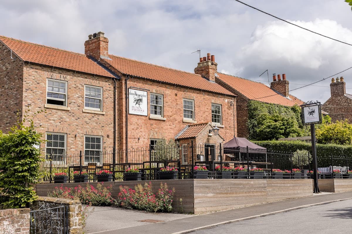 Brick building with a sign for "Plum & Partridge," outdoor seating, and flower beds in a quaint village setting.