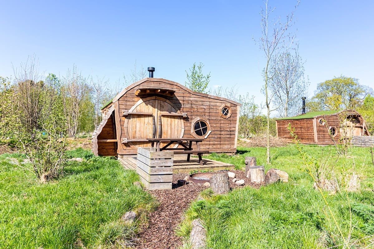 Wooden cabin with round windows, surrounded by greenery and trees, featuring a fire pit and rustic outdoor setting.