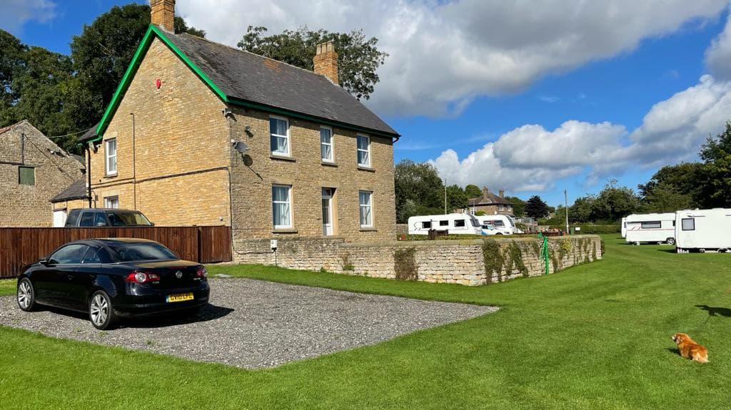 Stone building with green accents, parked car, grassy area, and caravans in the background under a blue sky.