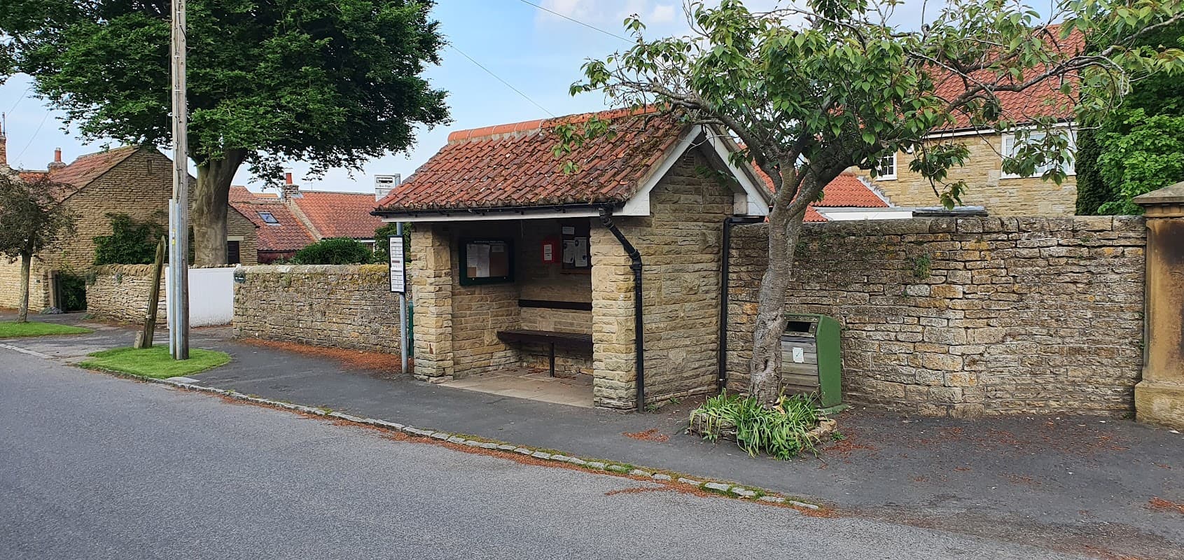 Hutton Buscel bus shelter with a stone wall, bench, and nearby greenery along a quiet street in Yorkshire.