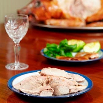 Sliced meat on a plate with a crystal glass and garnishes, with a whole roasted pig in the background.