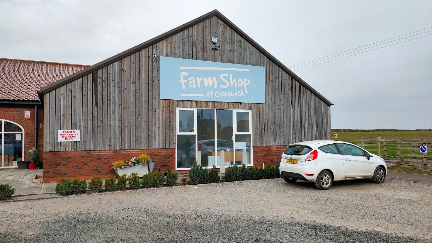 Wooden-clad farm shop with large windows, a white car parked outside, and greenery lining the entrance.