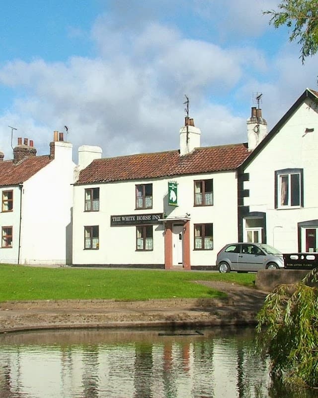 The White Horse Inn with a green sign, white walls, and a car parked beside a pond under a blue sky.