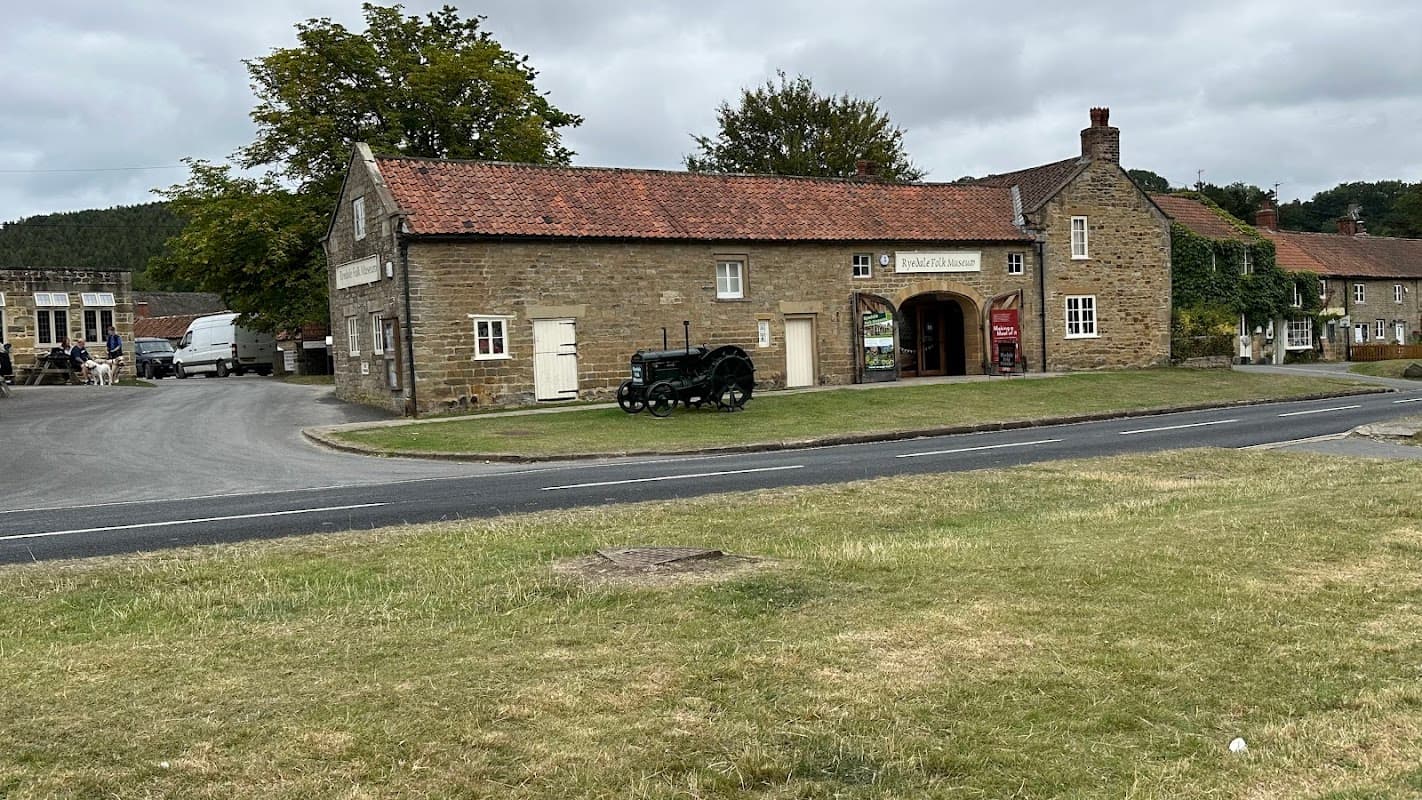 Historic stone building with a red-tiled roof, parked tractor, and nearby vehicles in a rural village setting.