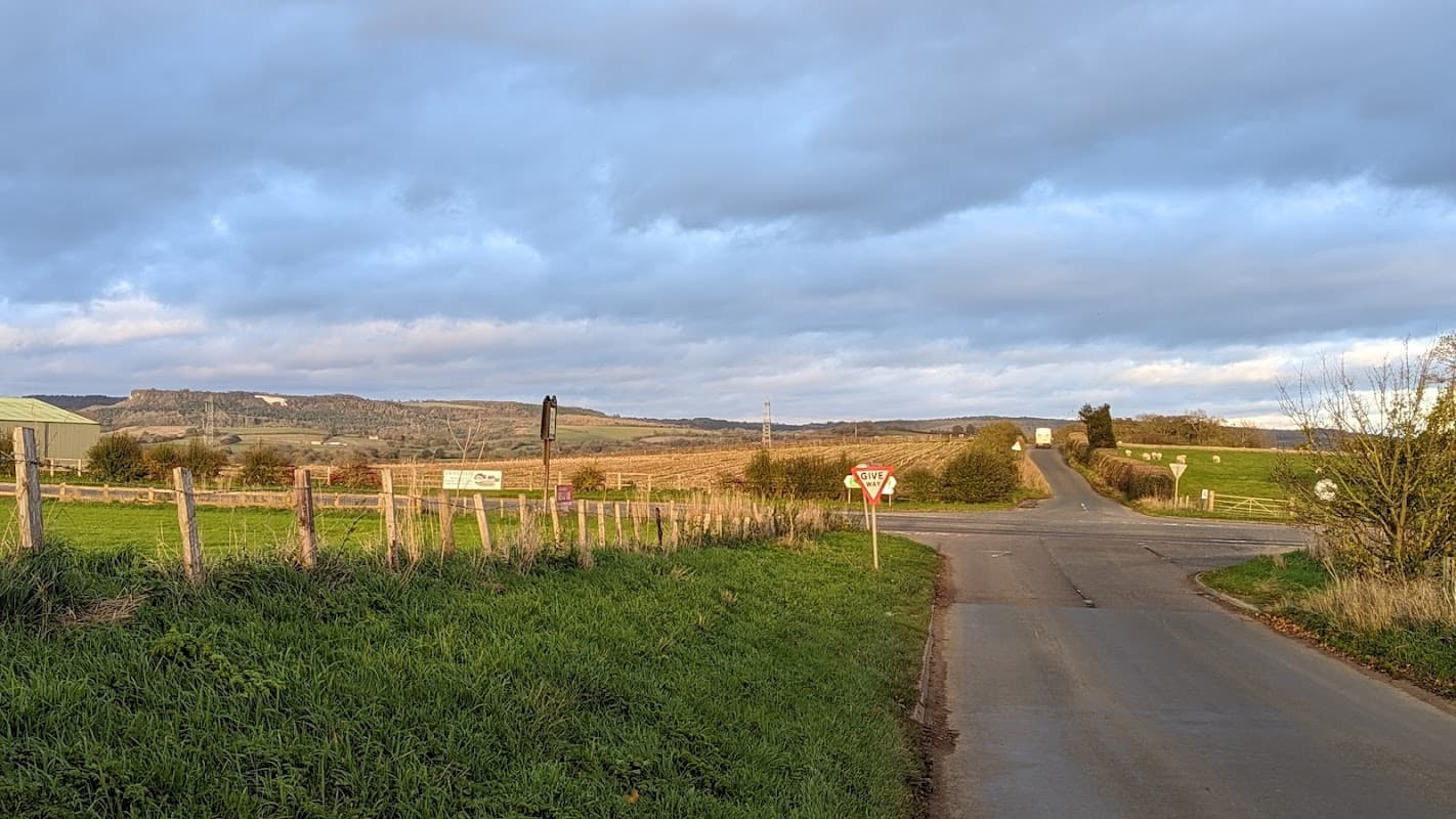 Rural road intersection in Hutton Sessay, with fields, a signpost, and distant hills under a cloudy sky.
