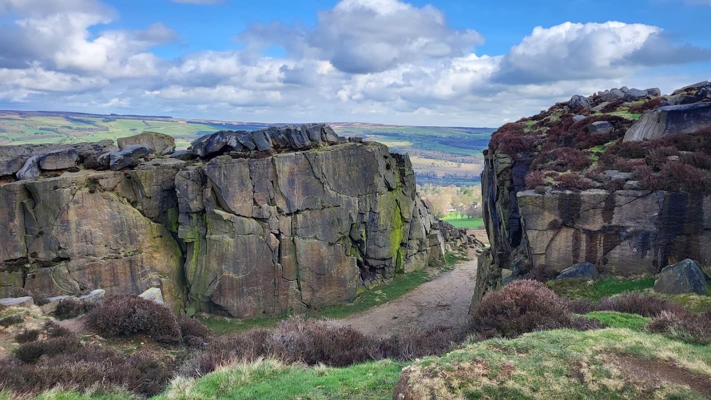 Rocky cliffs and grassy terrain under a blue sky with fluffy clouds, overlooking a scenic landscape in Ilkley Moor.