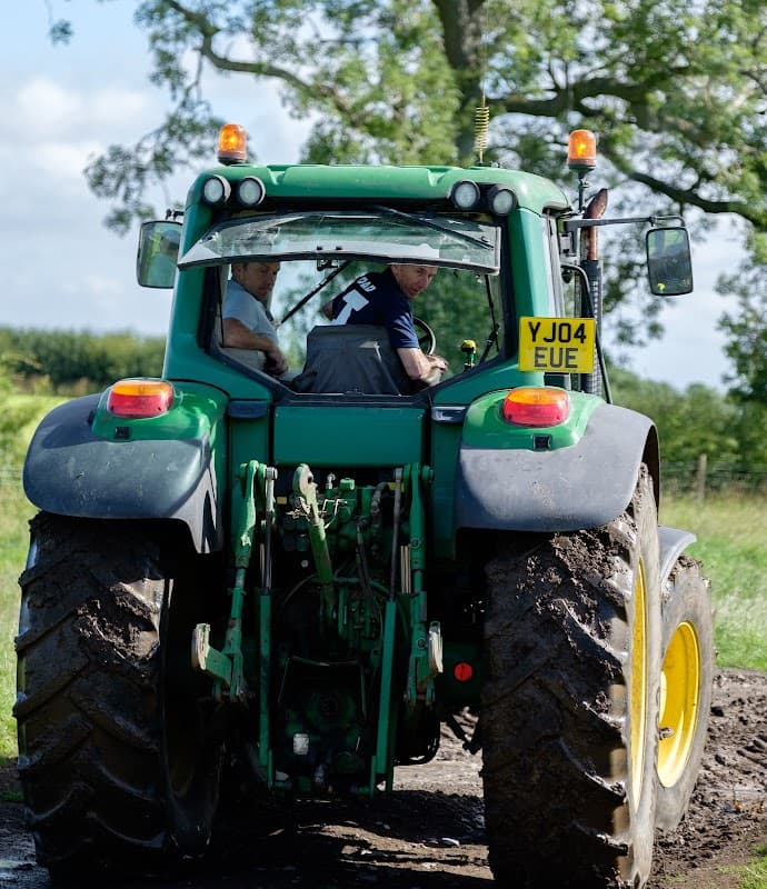 Two men in a green tractor on a dirt path, surrounded by lush greenery and blue skies in Yorkshire.