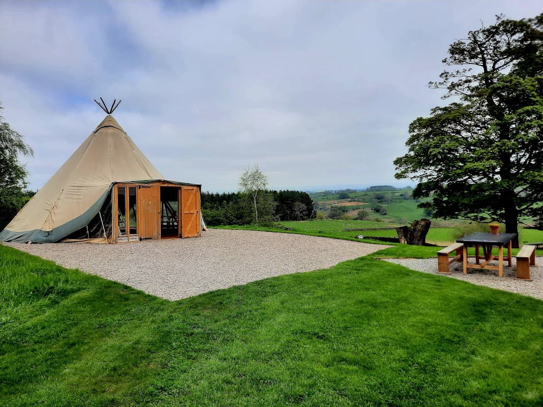 A large teepee tent on a gravel area, surrounded by lush green grass and trees, with rolling hills in the background.