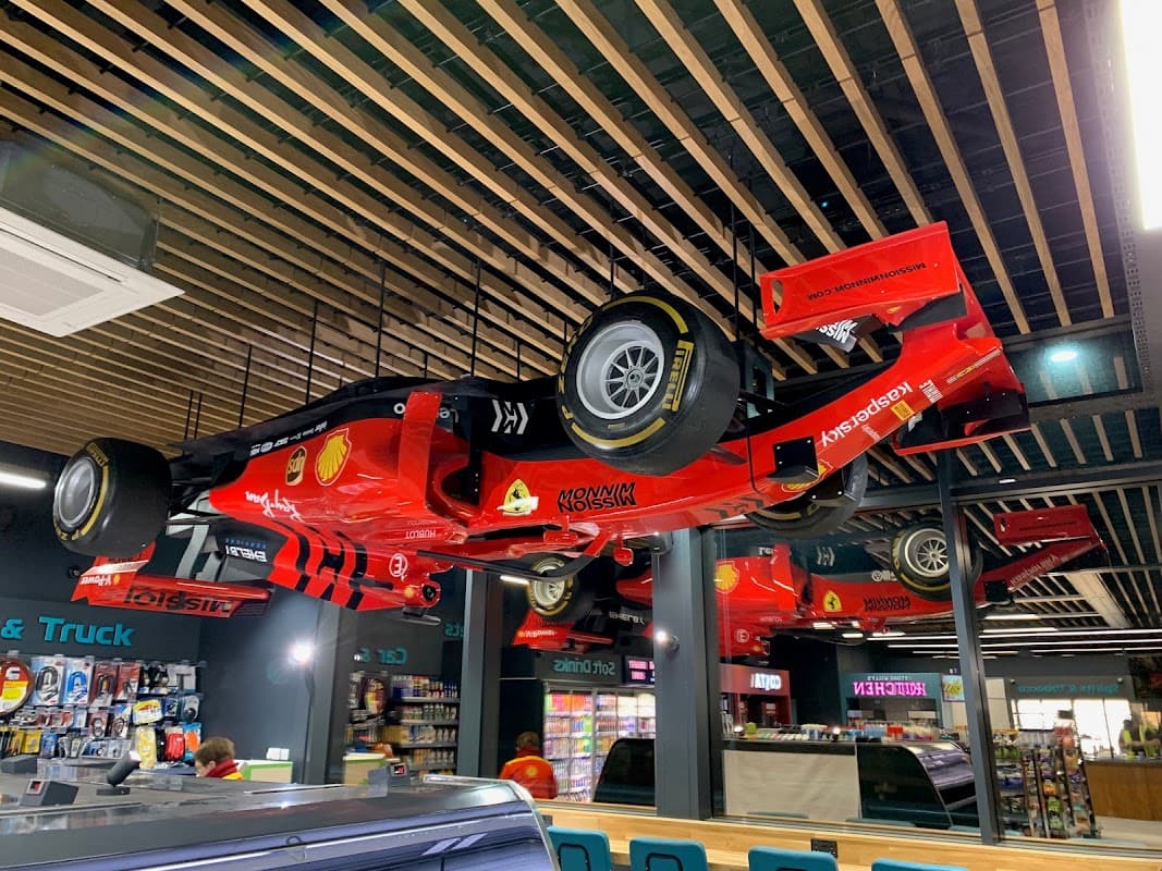 Ceiling-mounted red race cars with Shell logos, surrounded by shelves of snacks and drinks in a shop interior.