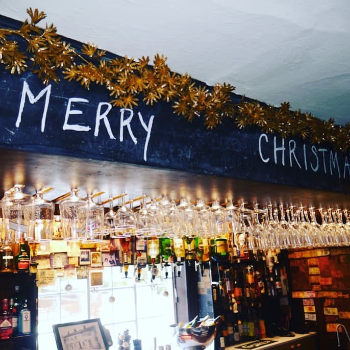 Bar interior with festive decorations, "MERRY CHRISTMAS" written on a chalkboard, and rows of hanging glasses.