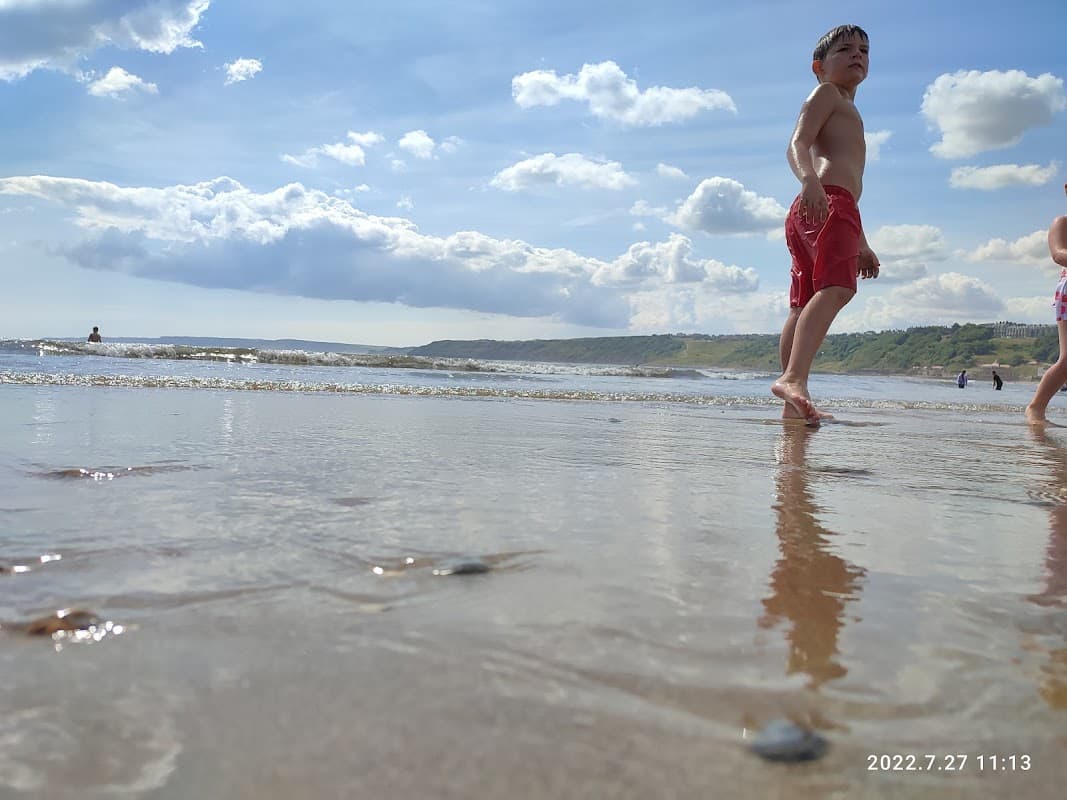 A child in red swim trunks walks along a sandy beach with gentle waves and a cloudy sky in the background.
