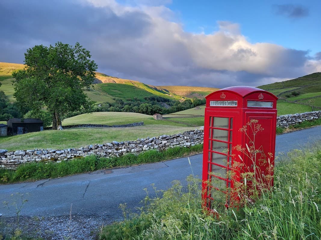 Red telephone box beside a winding road, with green hills and a tree in the background under a cloudy sky.