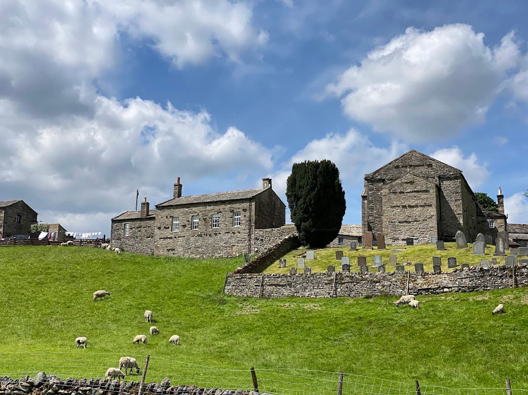 Stone buildings and a graveyard on a grassy hill, with sheep grazing under a partly cloudy sky in Keld, North Yorkshire.