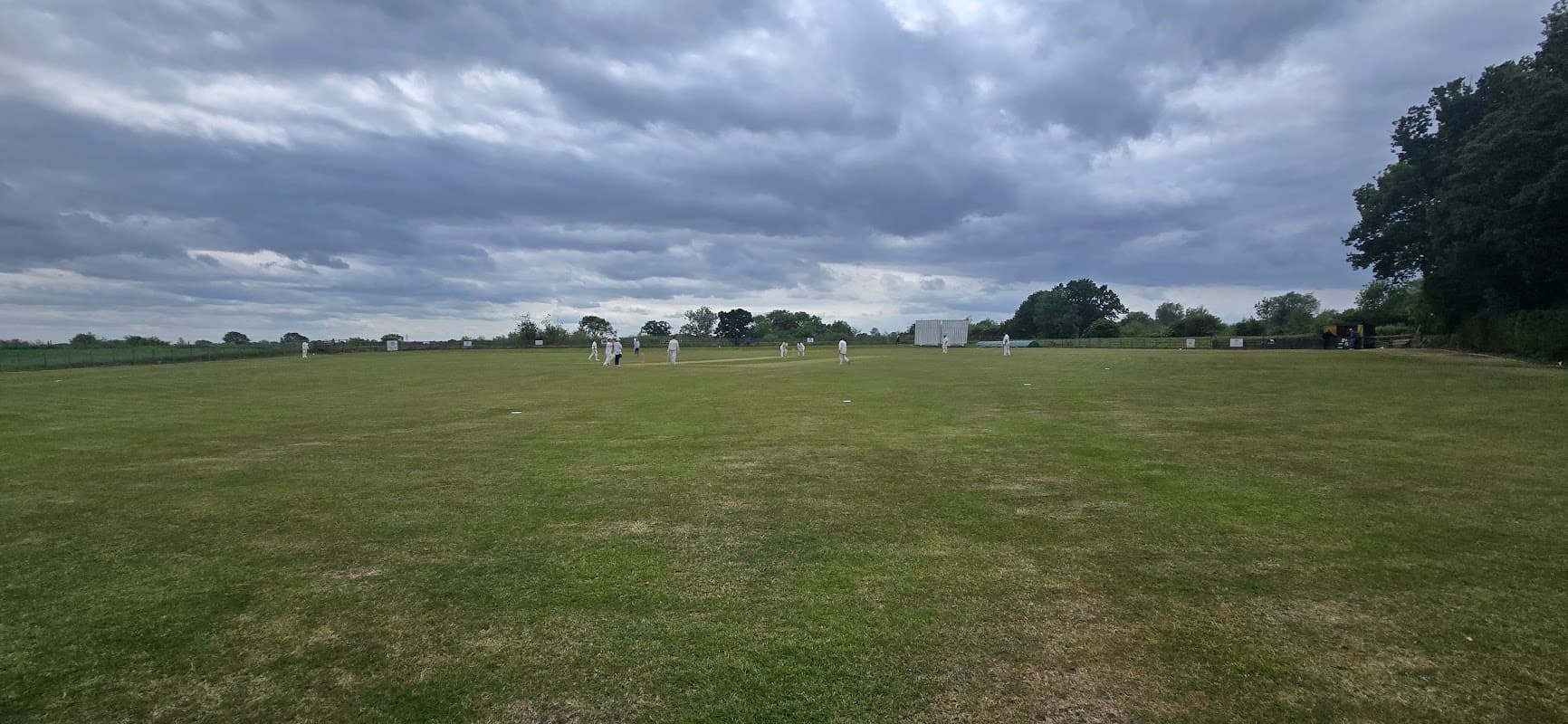 Cricket match in progress on a green field under a cloudy sky at Kelfield Cricket Club, North Yorkshire.