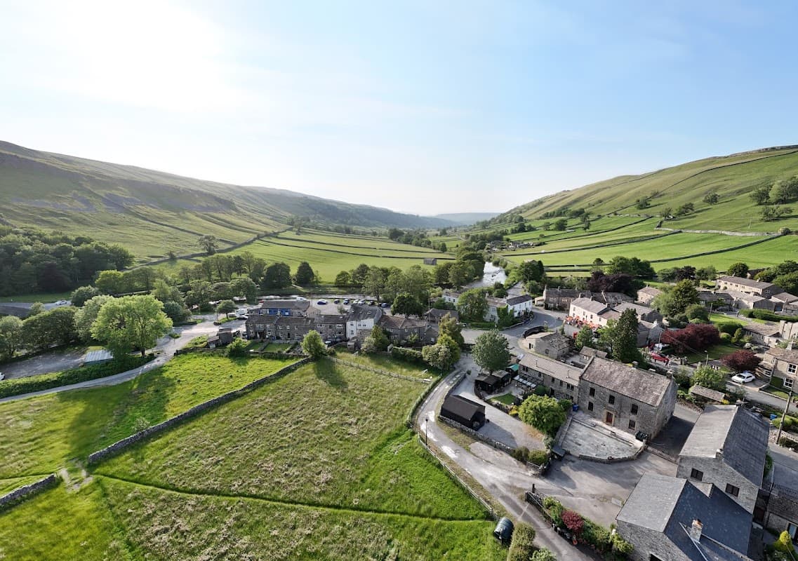 Aerial view of Kettlewell village, lush green hills, stone buildings, and winding river under a clear blue sky.