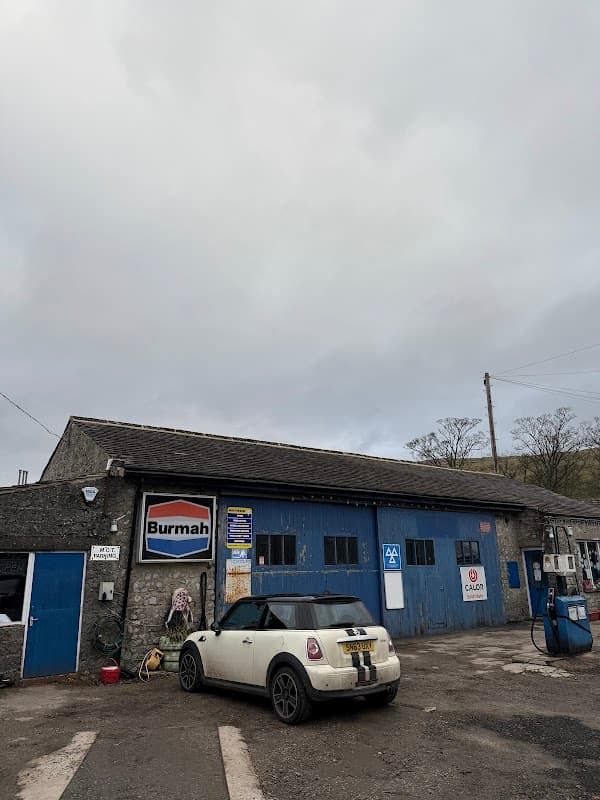 Kettlewell Garage with blue doors, a Burmah sign, and a white Mini car parked outside under a cloudy sky.