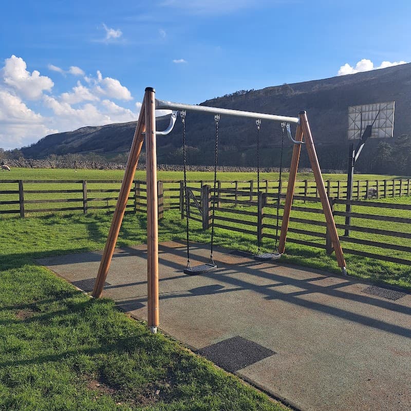 Wooden swingset in a grassy playground, surrounded by hills and a wooden fence under a blue sky with clouds.