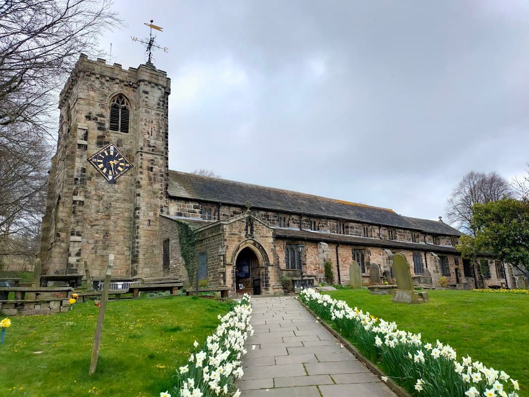 St. Andrew's Church with a stone tower, clock, and daffodils lining the path in a tranquil Yorkshire setting.