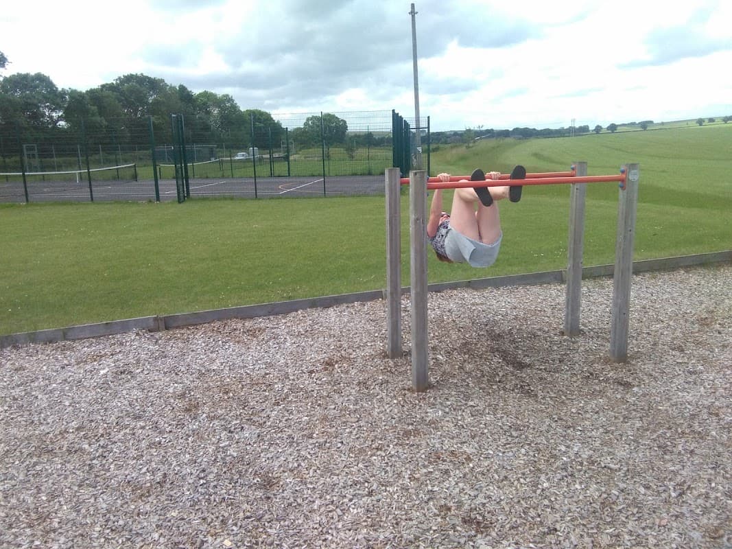 A child hangs upside down on a horizontal bar in a grassy field with a tennis court in the background.