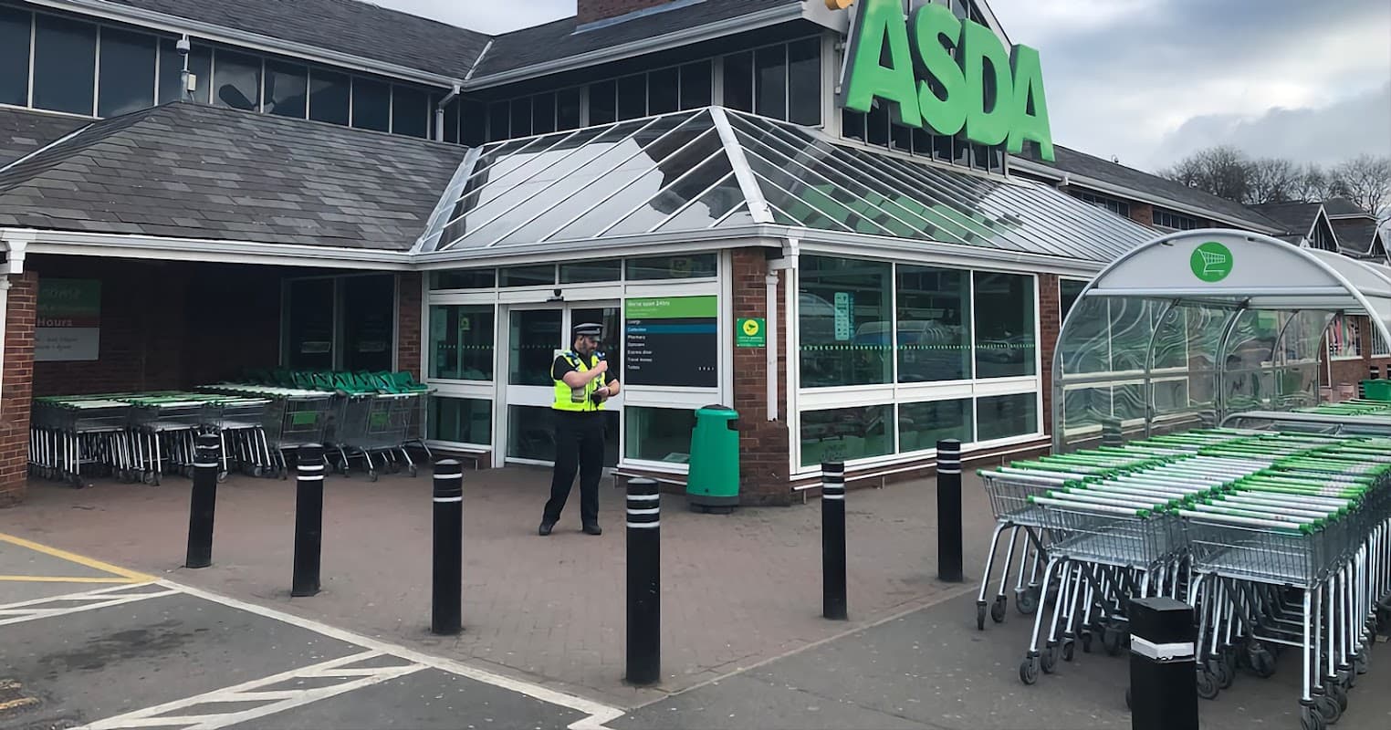 ASDA Pharmacy storefront with shopping carts outside and a security guard standing at the entrance.