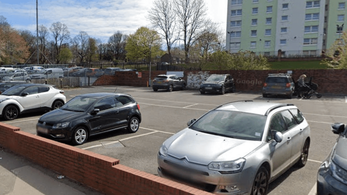 Cars parked in a lot near a residential building, with trees and a clear sky in the background.