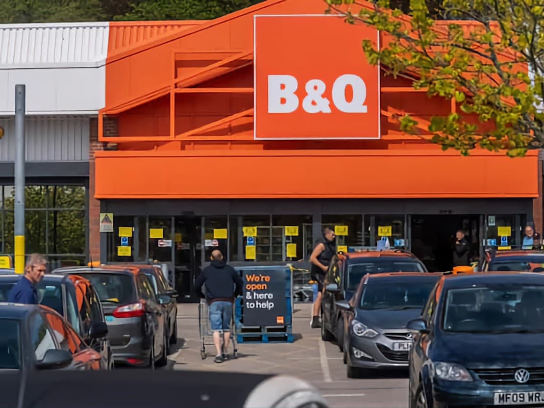 B&Q store entrance with customers, parked cars, and a sign stating "We're open & here to help."