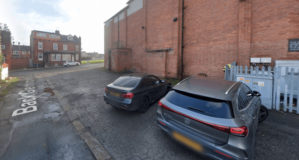 Two parked cars beside a brick wall and a gated area, with a street sign visible in the foreground.