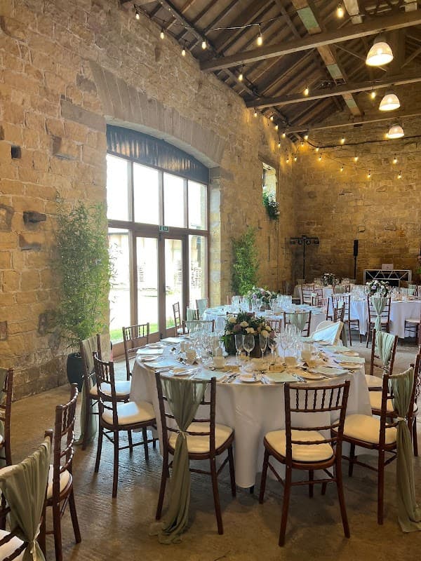 Elegant dining setup with round tables, white tablecloths, and greenery in a rustic stone venue with hanging lights.