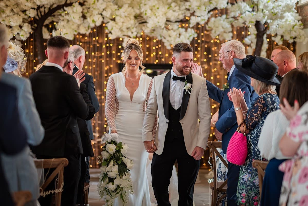 Bride and groom walk hand in hand down an aisle adorned with flowers and fairy lights, surrounded by applauding guests.