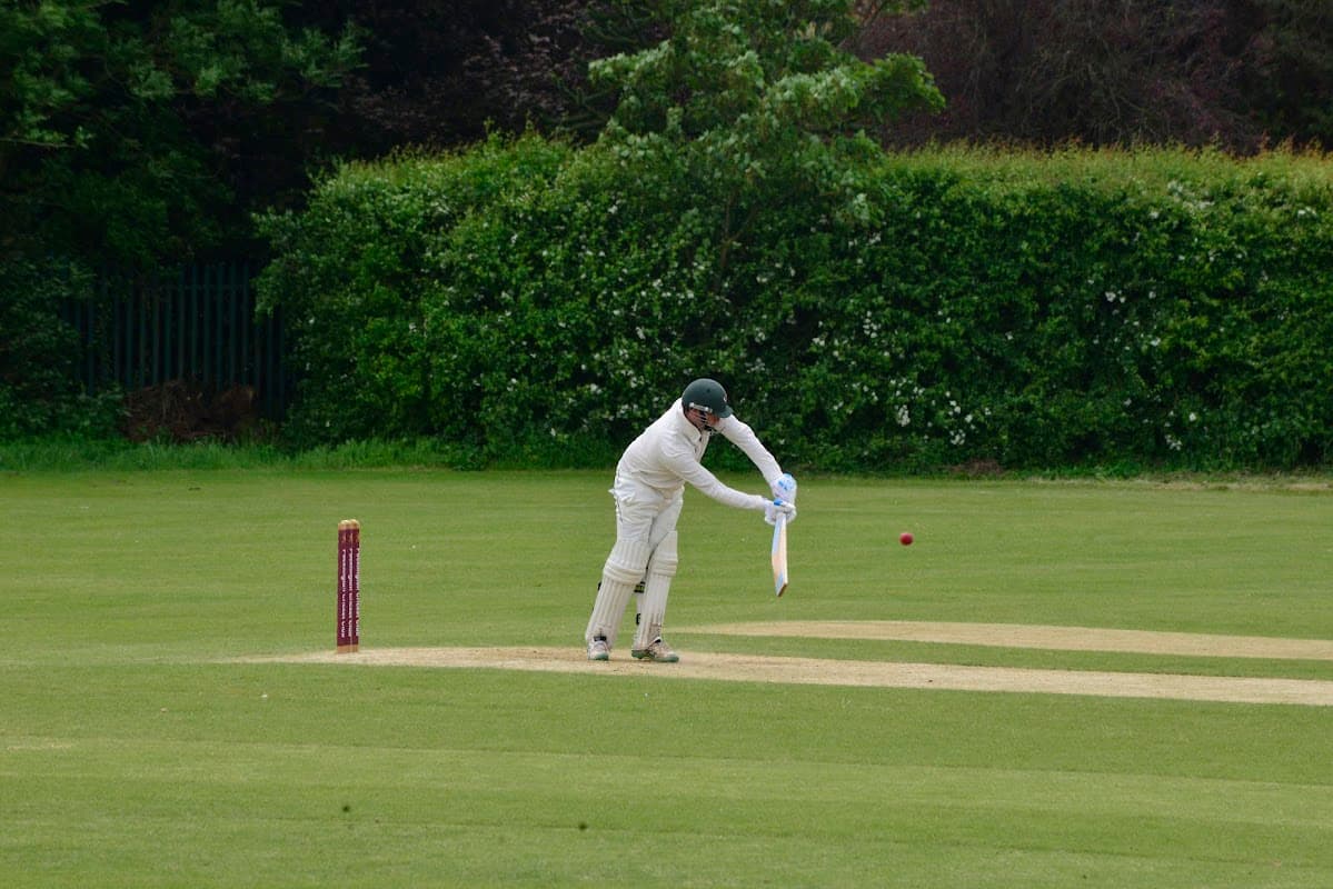 Cricketer in white uniform batting on a green field with a red cricket ball and stumps in the background.