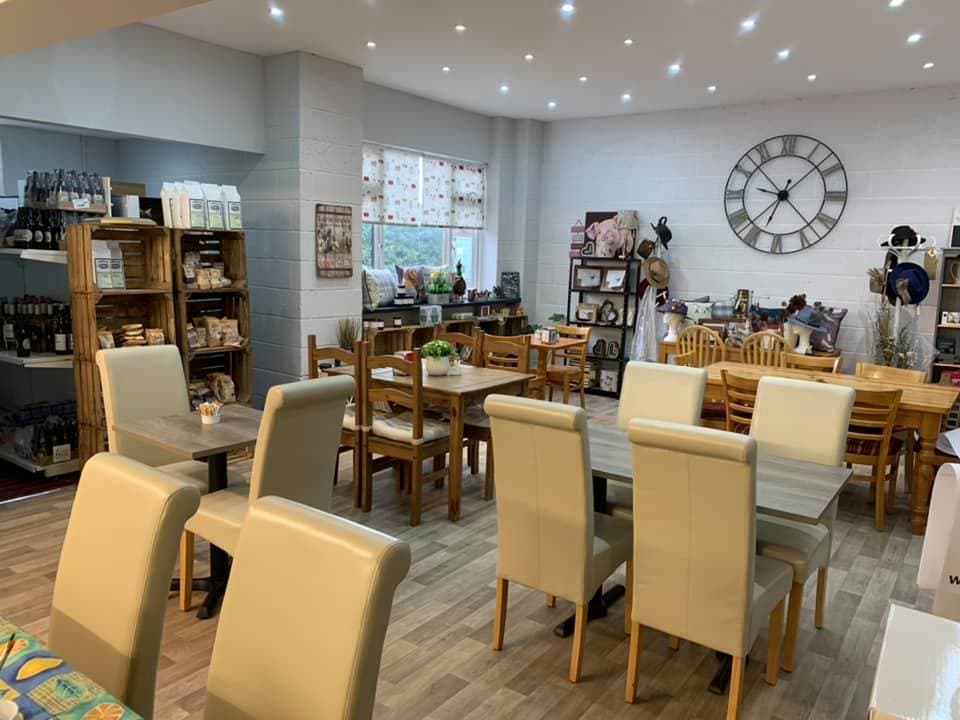 Cozy cafe interior with wooden tables, chairs, a large clock, and shelves displaying various products. Brightly lit space.