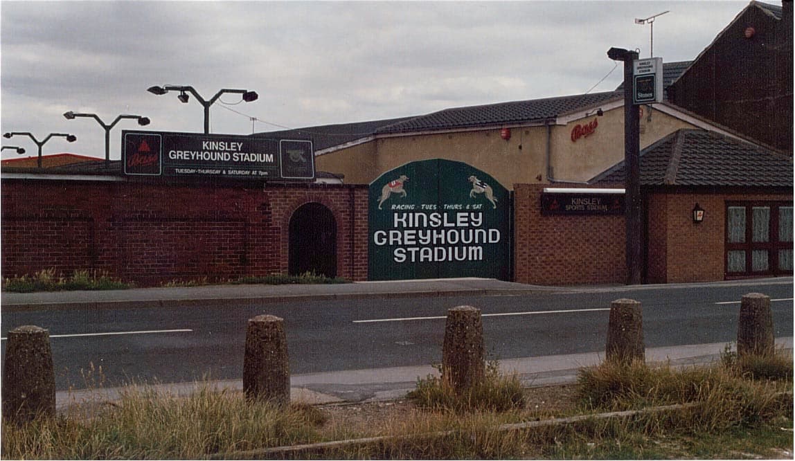 Kinsley Greyhound Stadium entrance with signage, brick walls, and a road in front. Overcast sky in the background.