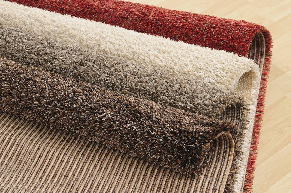 Four rolls of plush carpets in varying shades of red, cream, brown, and beige, displayed on a wooden floor.