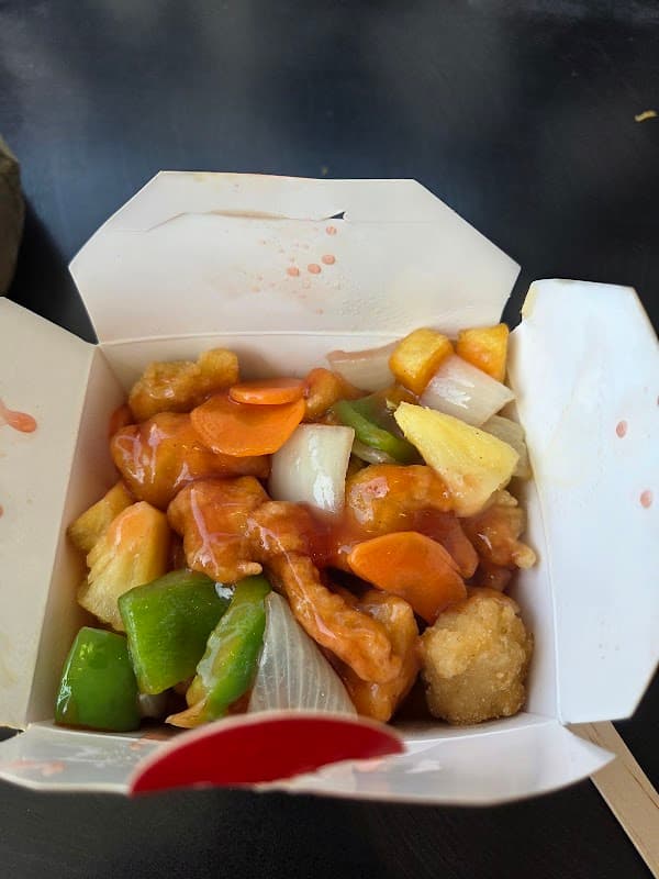 Takeout box filled with sweet and sour chicken, bell peppers, pineapple, and carrots in a tangy sauce.