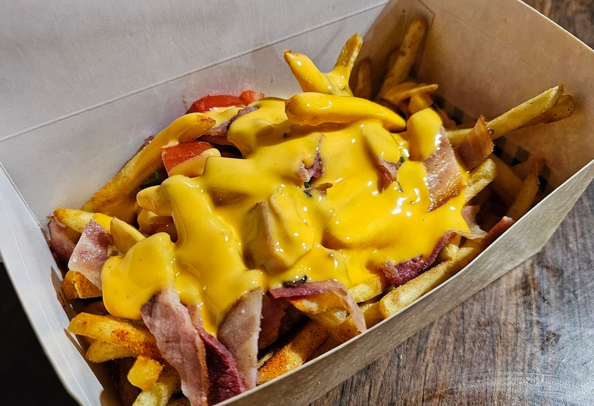 Fries topped with bacon, diced tomatoes, and melted cheese in a cardboard container.