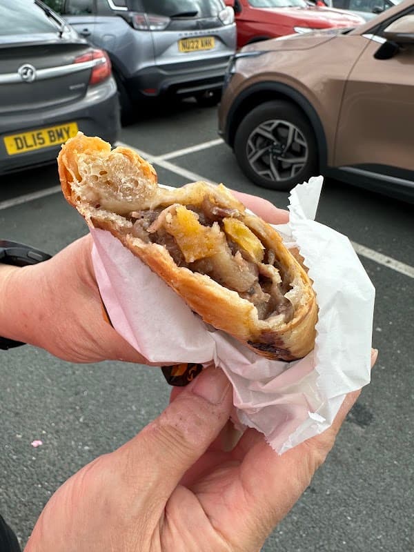 A hand holding a flaky pastry filled with meat and vegetables, with parked cars in the background.