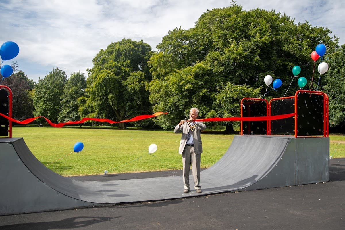 A man in a suit cuts a red ribbon at a skate park, surrounded by trees and balloons in a grassy area.