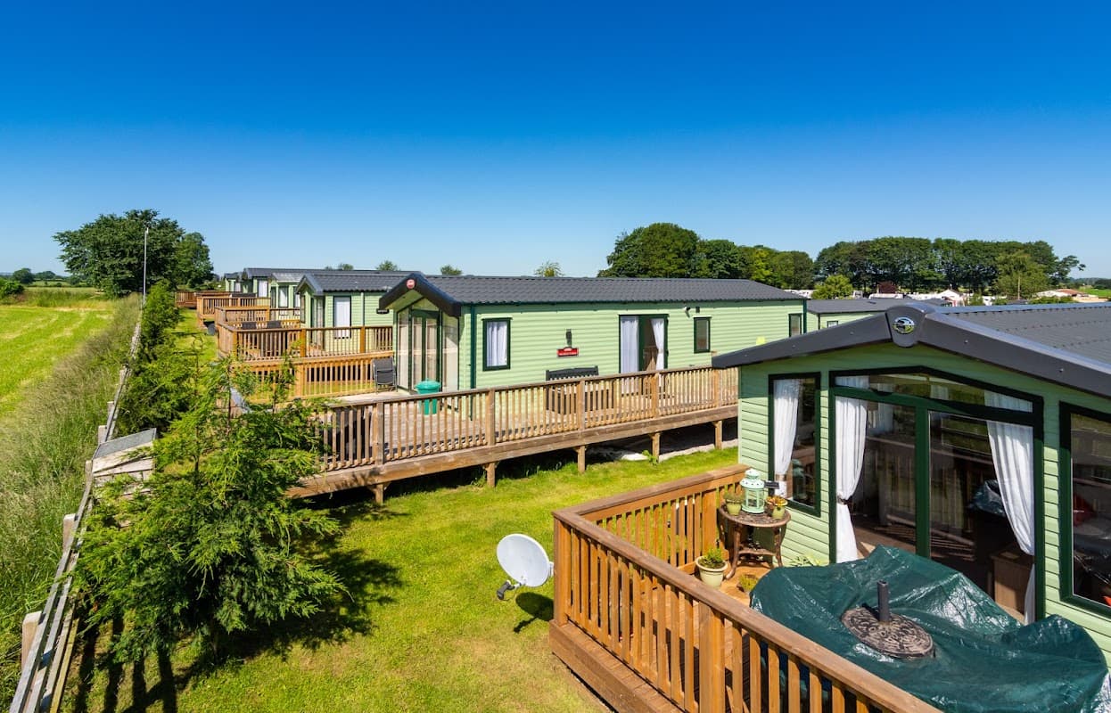 Lush green holiday park with cabins on wooden decks, surrounded by fields under a clear blue sky.